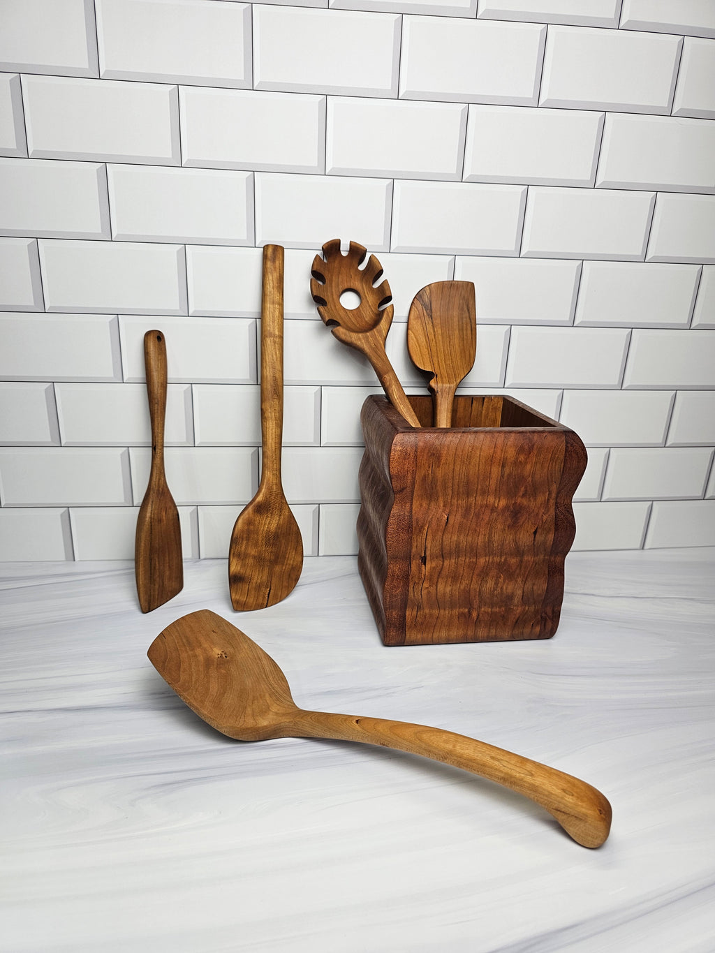 Set of wooden kitchen utensils including a spaghetti spoon and spatulas on a white tiled background