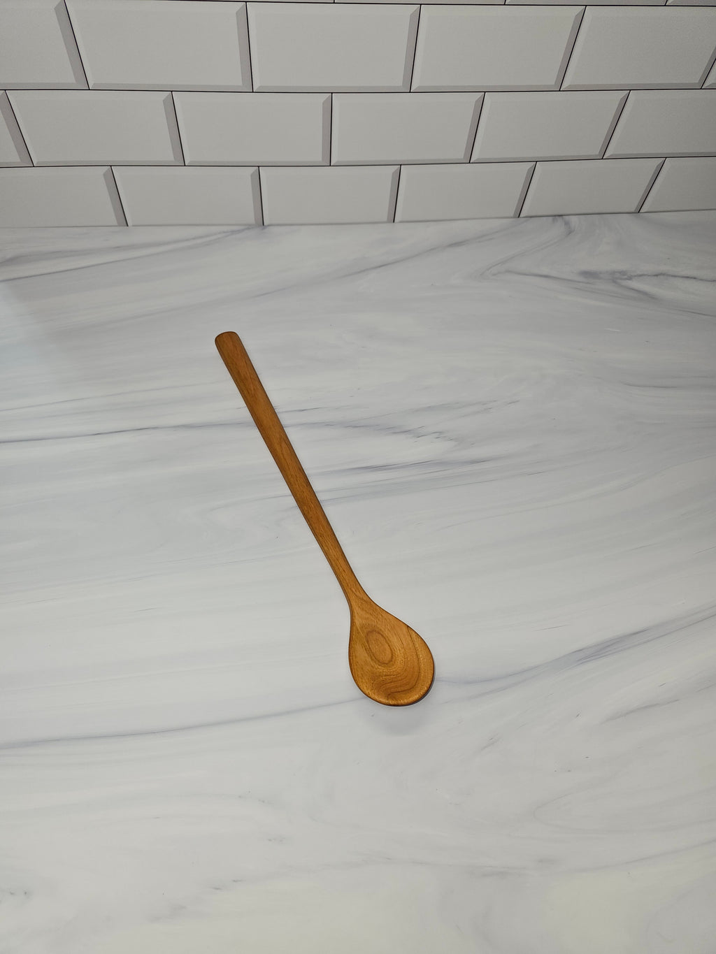 A single wooden spoon with a long handle and oval bowl, shown standing on a white marble-look countertop with a white subway tile backsplash behind it.
