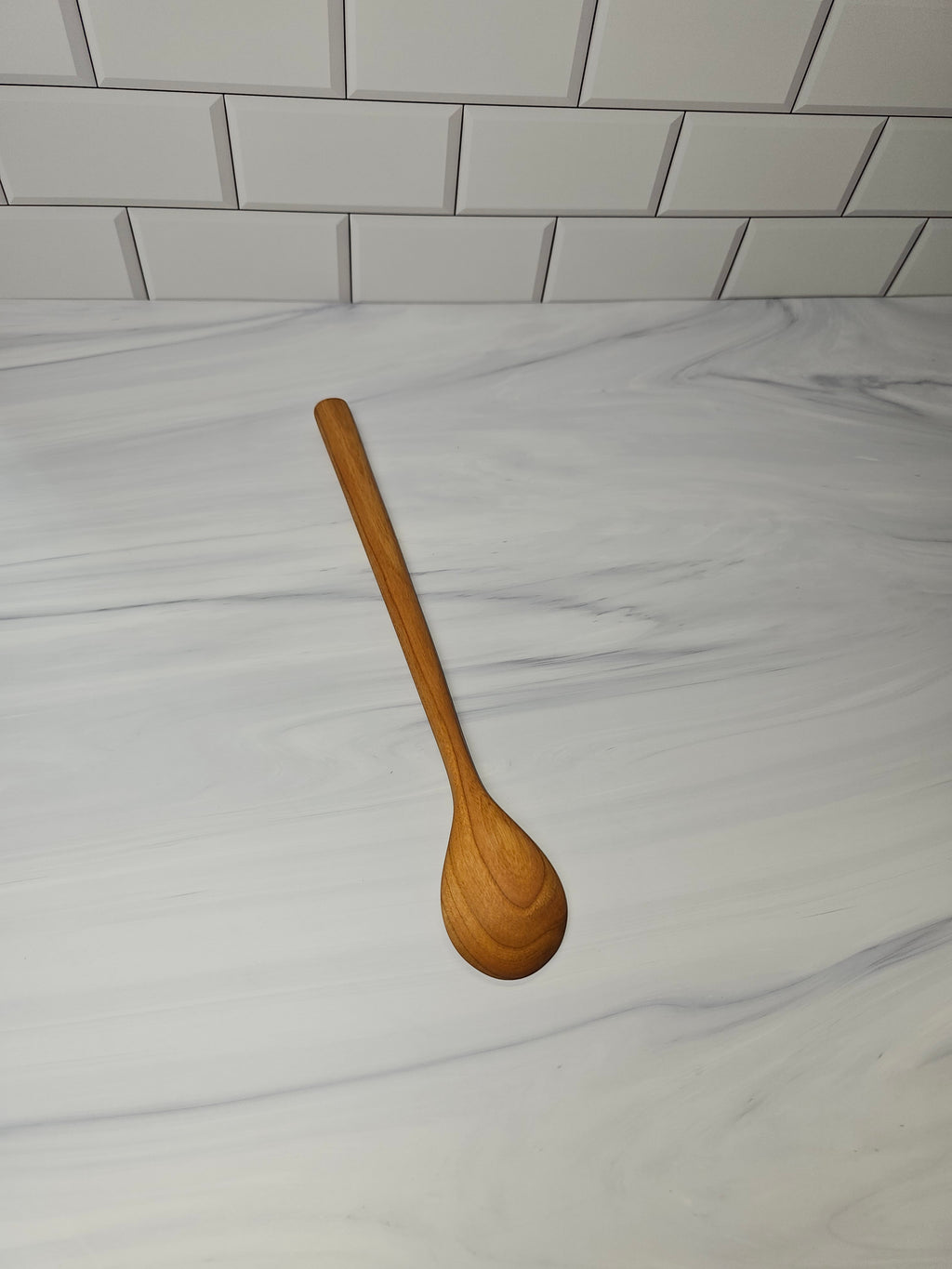 A single wooden spoon with a long handle and rounded bowl, shown diagonally on a white marble countertop with a white subway tile backsplash.