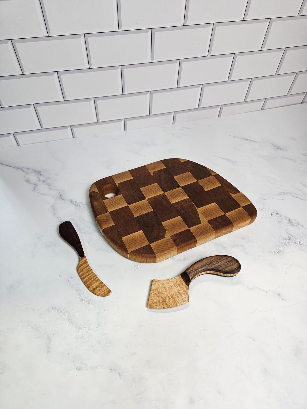 A wooden cutting board with a checkerboard pattern, accompanied by two matching wooden utensils, arranged on a white marble surface next to a white subway tile wall.