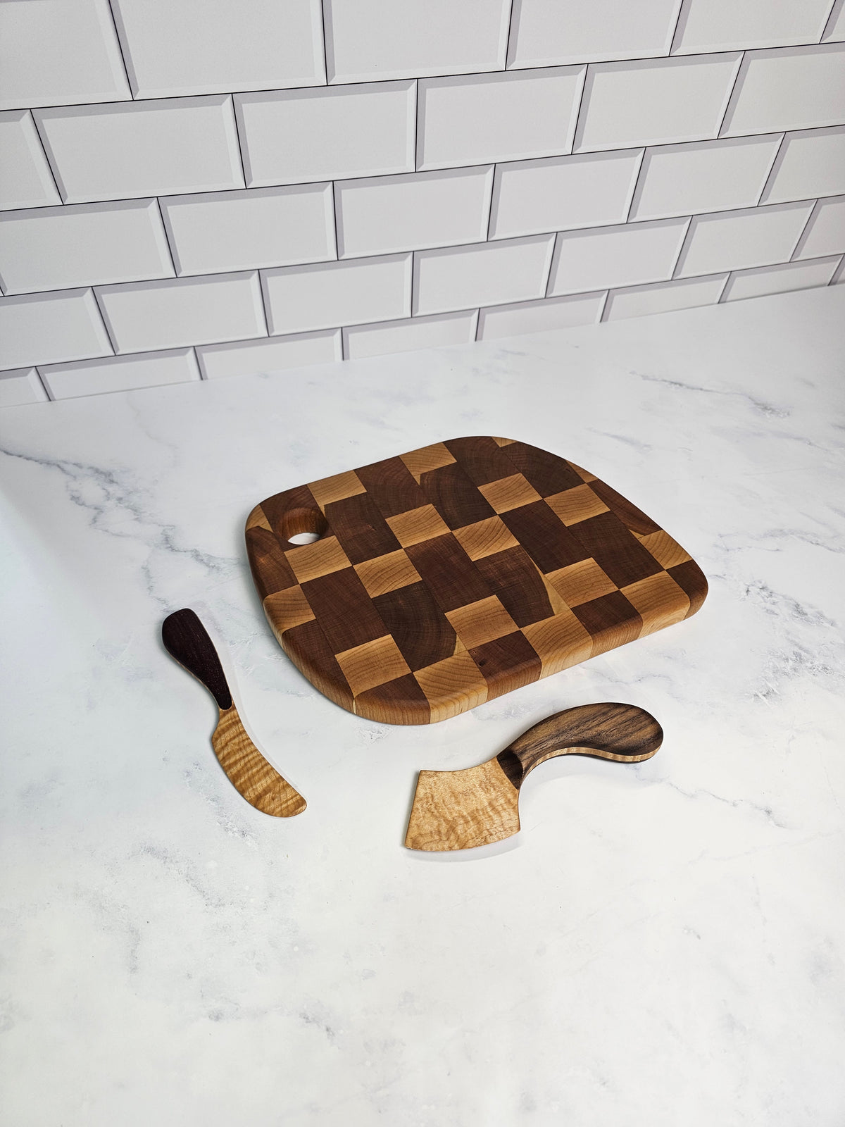 A wooden cutting board with a checkerboard pattern, accompanied by two matching wooden utensils, arranged on a white marble surface next to a white subway tile wall.